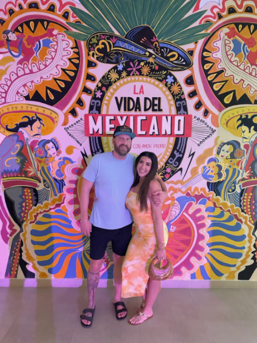 Travis and Chrissy posing in front of a colorful La Vida Del Mexicano mural.