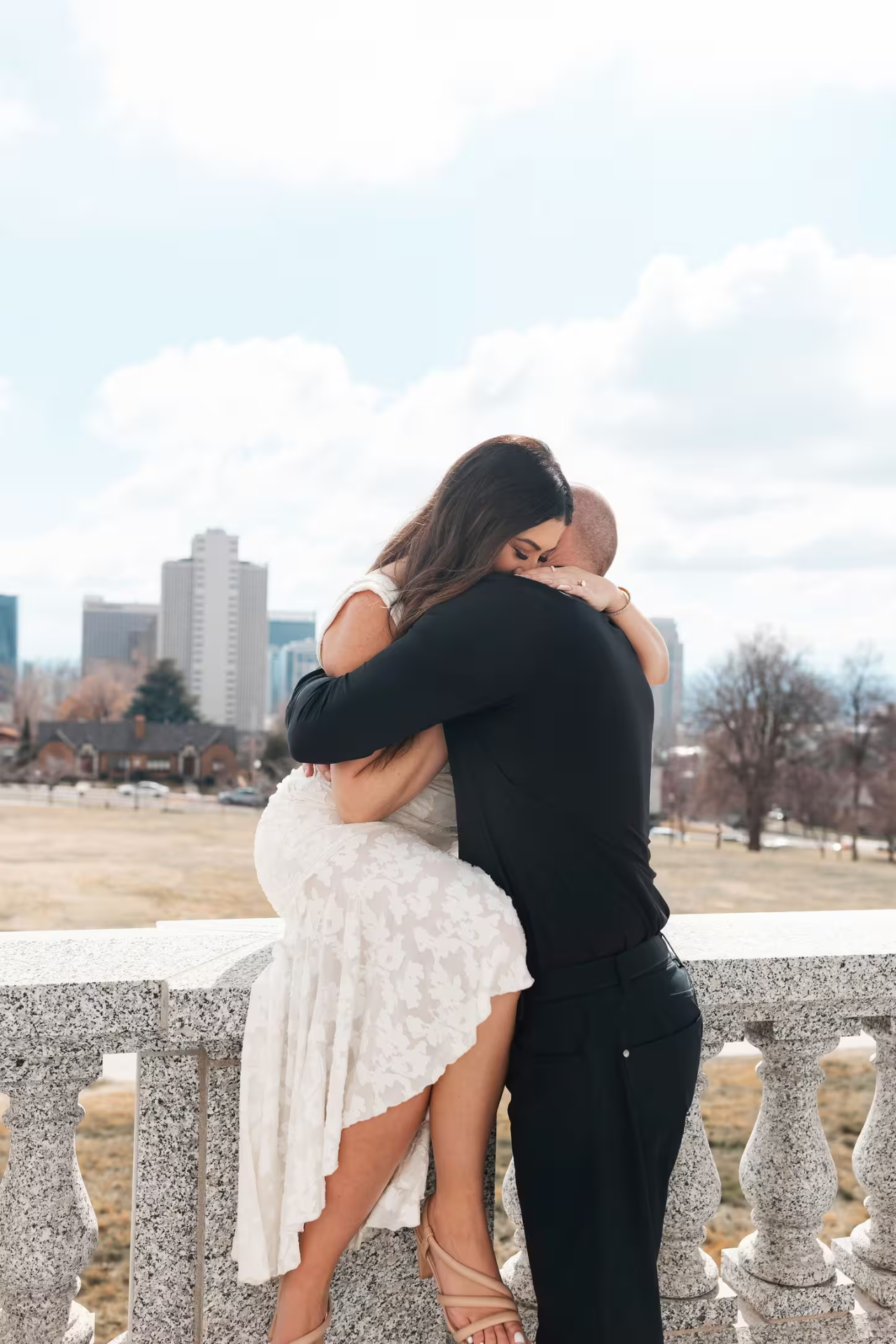 Travis hugging Chrissy as she sits on a stone railing outdoors.